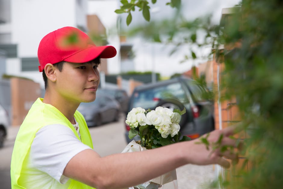 A young male florist wearing a red cap and a high-visibility yellow vest arranging a bouquet of fresh white hydrangeas in an outdoor setting. The arrangement is being carefully positioned near a green
