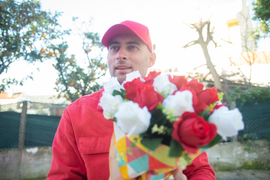 A delivery person dressed in a red uniform and matching cap holds a colorful floral bouquet featuring red and white roses, with hints of green foliage, wrapped in a patterned paper. The bouquet is pos
