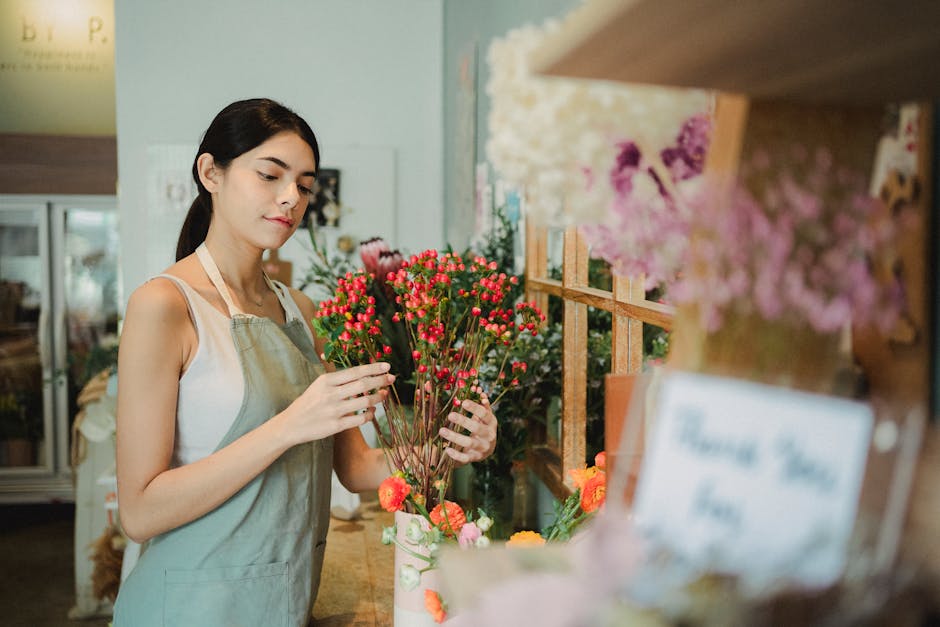 A young female florist with dark hair, wearing a beige apron over a white top, is carefully arranging a bouquet of small, bright pink and red flowers with green stems. She is standing behind a wooden 