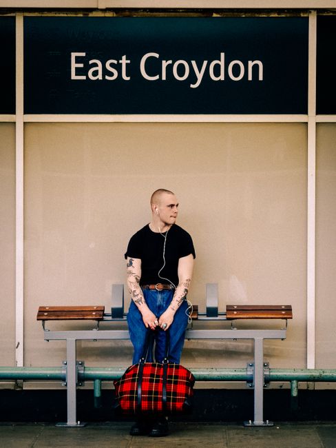 A young man with a shaved head, wearing a black t-shirt and blue jeans, is sitting alone on a bench at East Croydon station. He has visible tattoos on his arms and is holding a red and black plaid bag