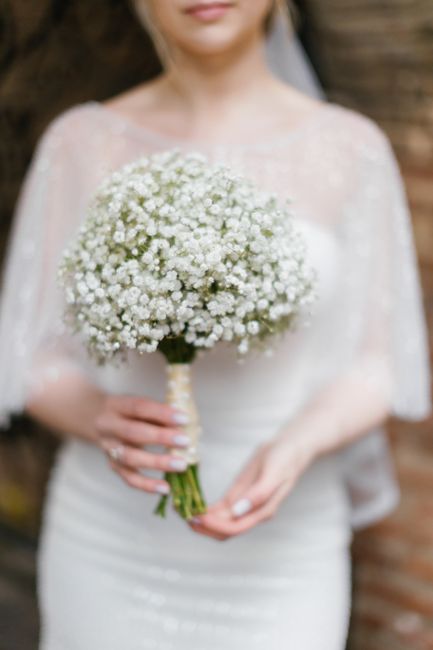 A bride in a delicate lace wedding gown holds a round bouquet of white baby's breath flowers with lush, tiny blooms and green stems wrapped in a cream ribbon. Her face is out of focus, emphasizing the