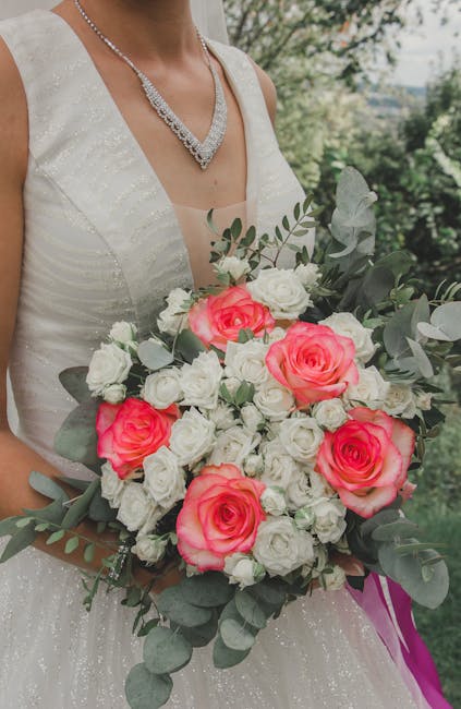 Close-up of a bride wearing a sleeveless, ivory wedding gown with subtle shimmer, holding a large floral bouquet outdoors. The bouquet features vibrant pink roses, white roses, and smaller white fille